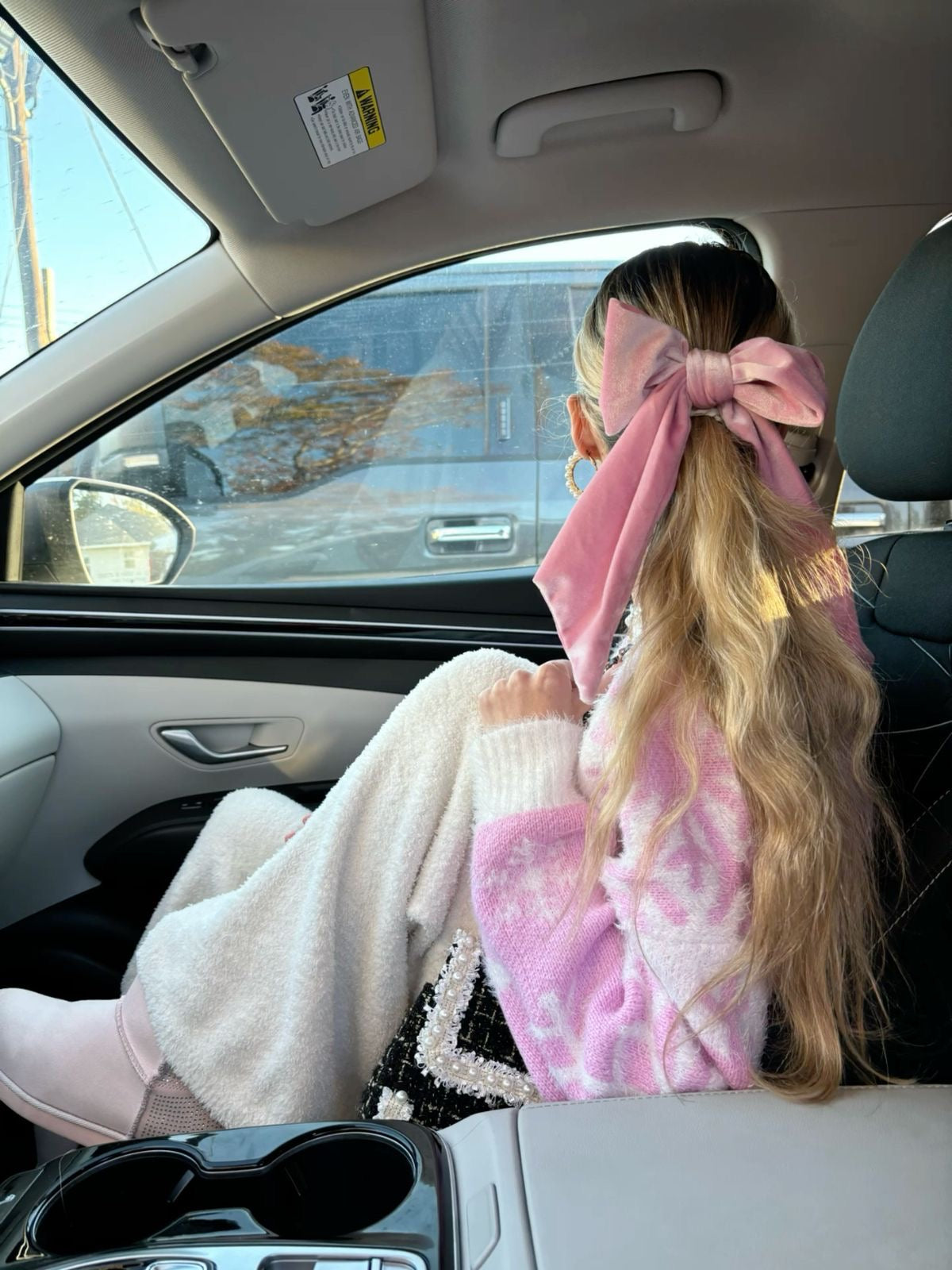 Person sitting in a car with a large pink bow in their hair, wearing a white sweater.