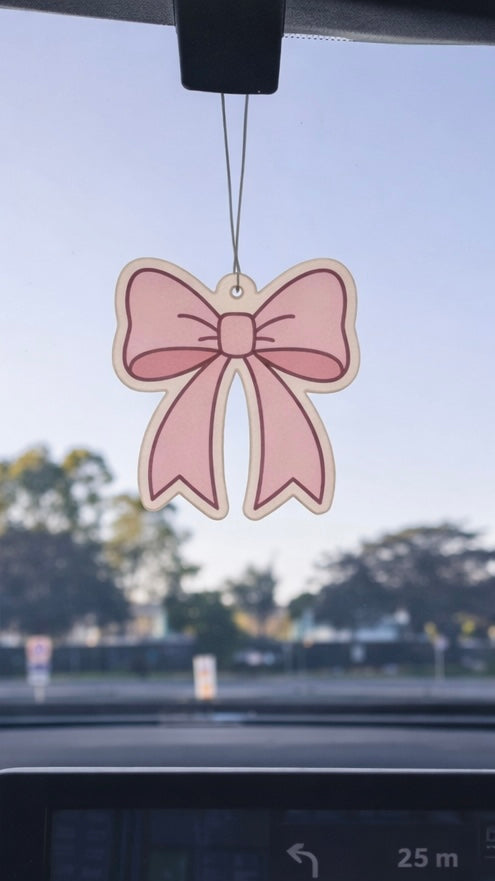 Pink bow-shaped air freshener hanging from a car's rearview mirror with a blurred street view in the background.